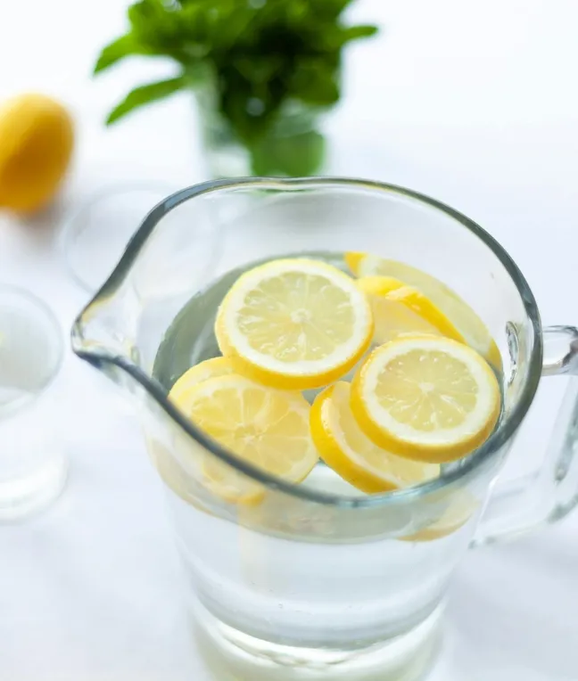 Ein Glaskrug mit Wasser und Zitronenscheiben, im Hintergrund frische Minze und eine Zitrone.